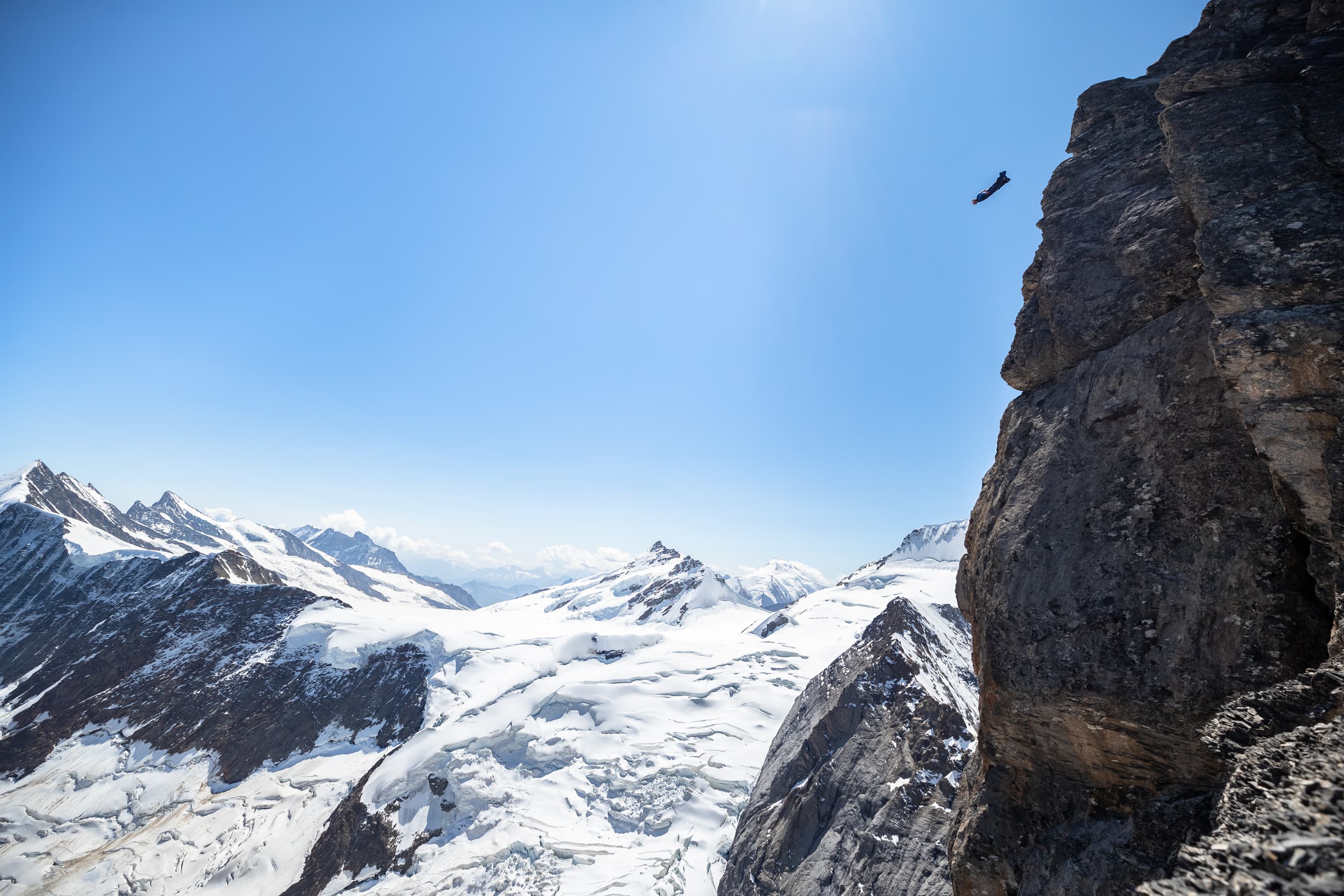 Ein BASE Jumper fliegt von einer Felswand über eine verschneite alpine Landschaft.