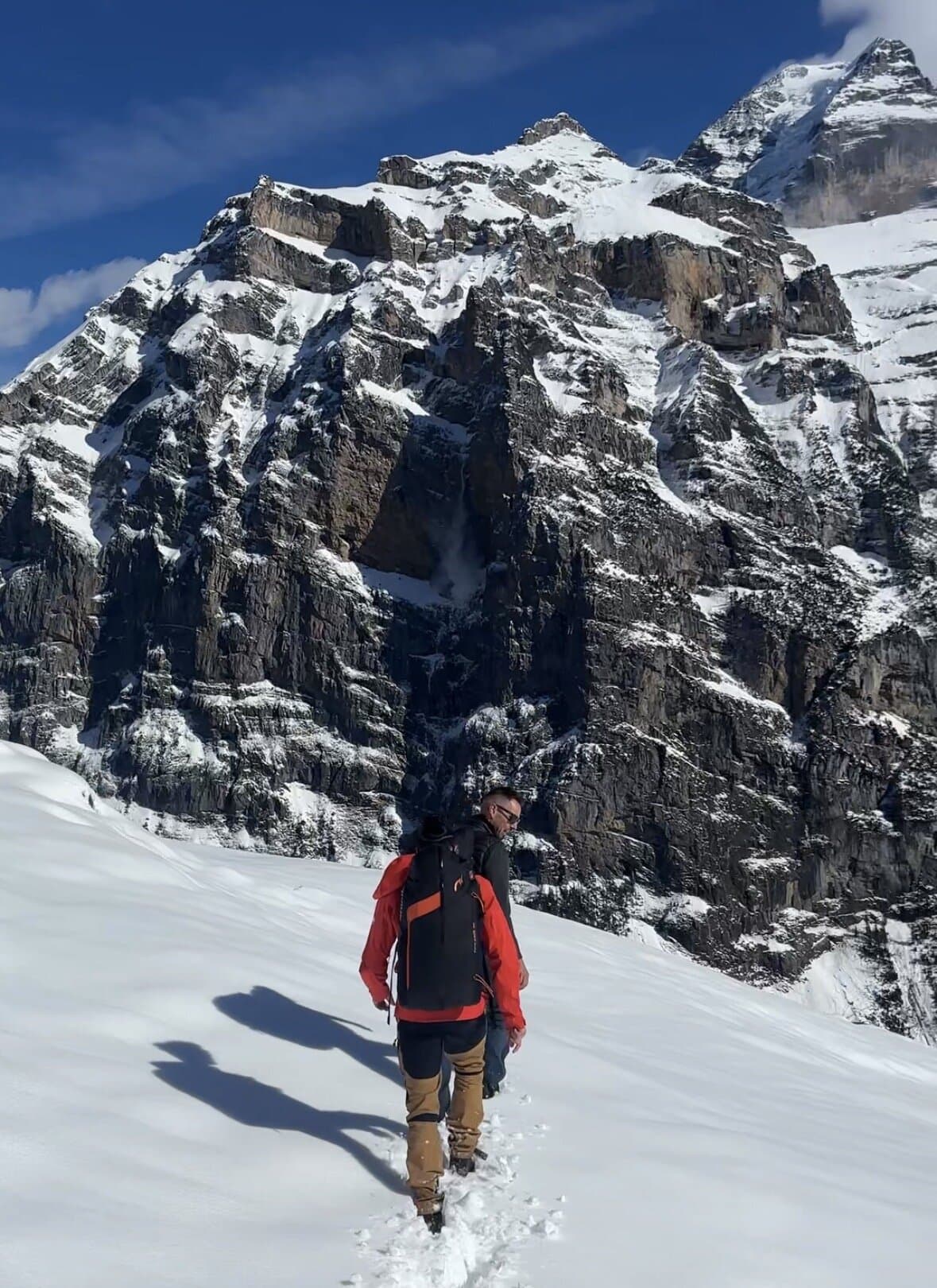 Person walking across snow below a dark alpine rock wall