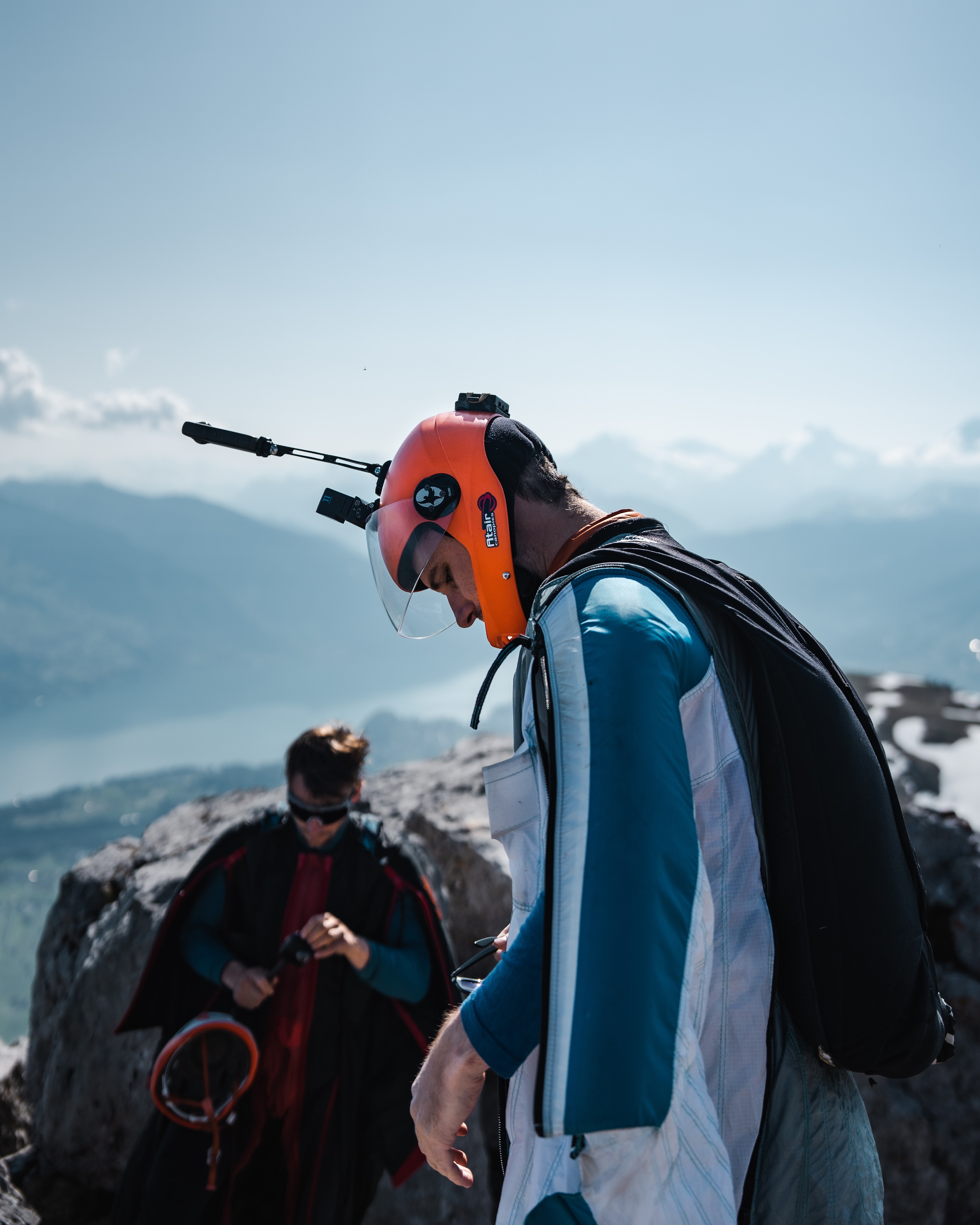 Ein BASE-Athlet bereitet sich an einem Berg auf einen Sprung vor. An seinem orangefarbenen Helm ist eine Actionkamera mit seitlich ausragender Halterung montiert. Im Hintergrund bereitet sich ein weiterer Athlet vor.